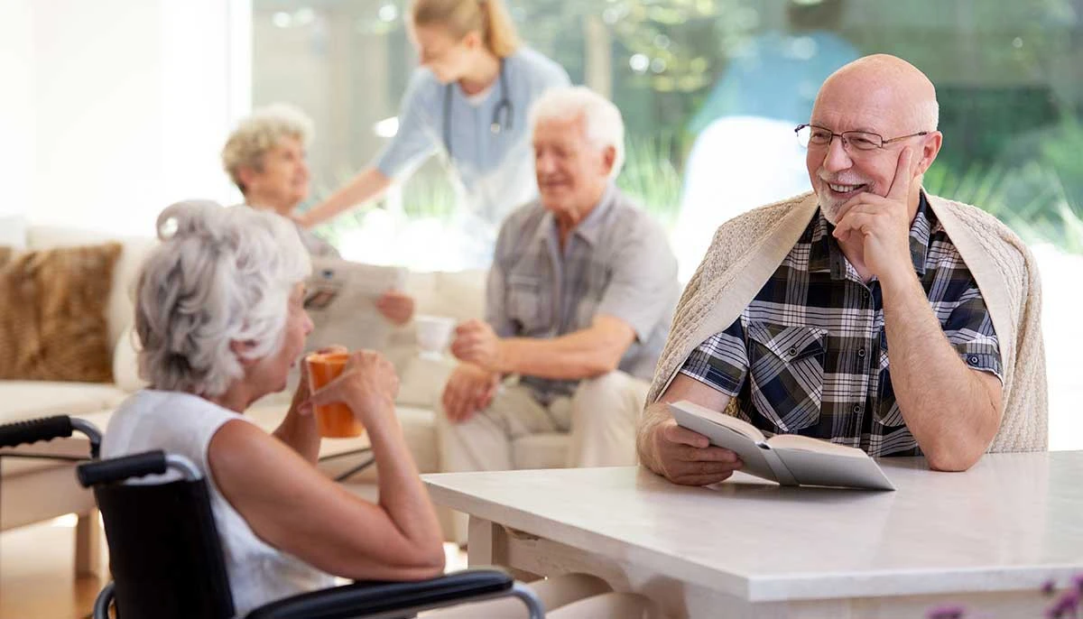 Group of seniors at a nursing home engaged in happy conversation while nurse checks in on patients