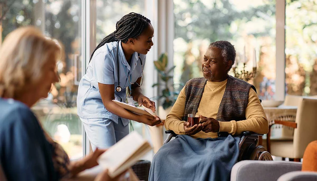 Elderly man in wheelchair drinking tea and talking to nurse in assisted living facility